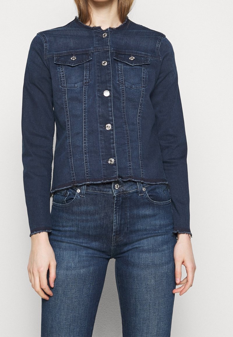 Chaqueta de mezclilla azul oscura con un diseño cortado, dos bolsillos en el pecho, botones a presión plateados y detalles deshilachados en el escote y en los puños.