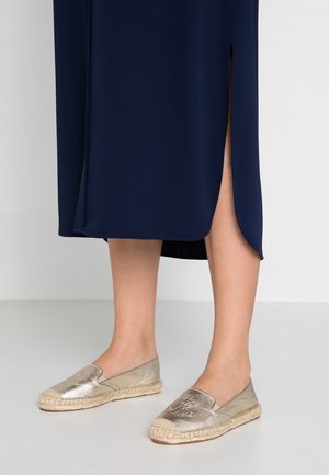 Femme portant des espadrilles slip-on métalliques avec semelles en corde et une jupe midi bleu marine fendue sur le côté, debout sur un sol clair.