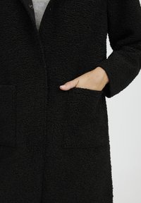 Fekete textúrájú kabát bolyhos anyagból, két elülső zsebbel és gombos záródással, sima háttér előtt.