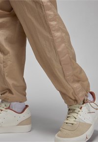 Beige träningsbyxor med en glansig rand som har texten "JORDAN", tillsammans med beige och krämfärgade sneakers och vita ankelsockor.