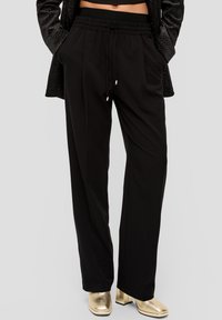 Close-up van zwarte wijd broek met elastische tailleband en trekkoord, gedragen met gouden tekstuur blokhak enkellaarsjes.