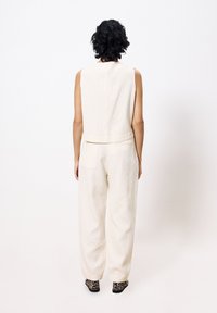 Mouwloze off-white jumpsuit van een gestructureerde stof, met een relaxte pasvorm en wijde benen. Bevat een achterzak en geprint zwarte schoenen.