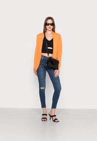 Blazer laranja, top preto cropped com recorte, jeans skinny azul desgastados, bolsa preta e sandálias de tiras pretas. Texturas suaves e cores vivas.