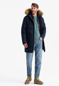 Parka imbottita blu marina con cappuccio foderato in pelliccia, due tasche frontali, indossata sopra una camicia teal e una flanella a quadri, abbinata a jeans chiari e stivali beige.