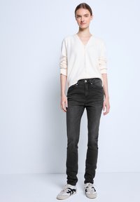 Jasno beżowy dziergany kardigan z dekoltem w serek i guzikami, zestawiony z ciemnoszarymi dopasowanymi dżinsami i szarymi trampkami z czarnymi akcentami.