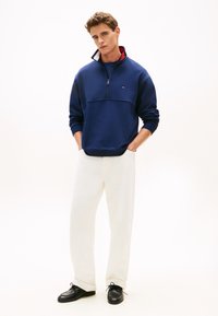 Μπλε σκούρο φούτερ με φερμουάρ στο μισό μέρος, με ριμπ μανσέτες και γιακά, συνδυασμένο με λευκό παντελόνι φαρδιάς γραμμής και μαύρα παπούτσια. Μινιμαλιστικός σχεδιασμός και απαλή υφή.