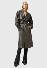 Trench coat in pelle verde oliva con vita cinturata, colletto ampio e maniche lunghe. Abbinato a pantaloni neri svasati e scarpe con punta.