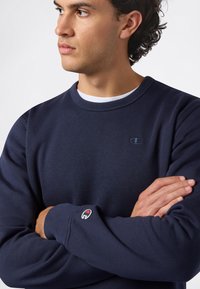 Sudadera azul marino de tela suave con cuello redondo, que presenta un pequeño logo en el pecho y un parche de la marca en la manga.