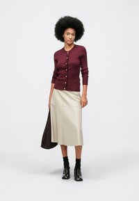 Mulher com cabelo afro a usar um cardigan bordeaux com botões, saia midi bege, meias pretas e sapatos castanhos-escuros, segurando uma bolsa castanha.
