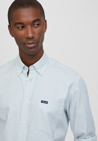 Camicia button-up a righe blu chiare con colletto a punta, dotata di una singola tasca sul petto e di un piccolo etichetta logo in navy. Tessuto di una consistenza liscia.