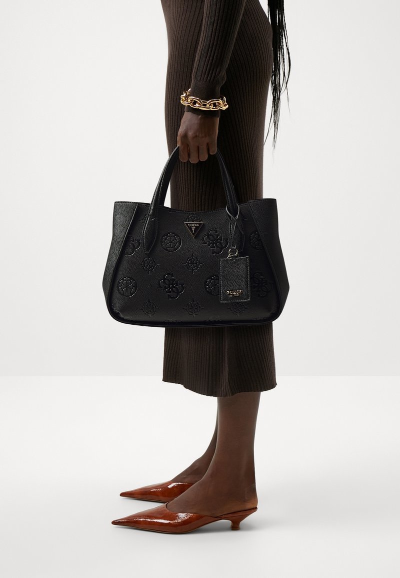 Femme portant une robe marron côtelée, des chaussures marron à talons bas pointus, un bracelet chaîne doré, tenant un sac à main embossé Guess noir avec étiquette.