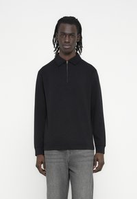 Μαύρο πουλόβερ με φερμουάρ στον γιακά και μακριά μανίκια, φτιαγμένο από λείο ύφασμα, συνδυασμένο με γκρι τζιν κανονικής γραμμής.