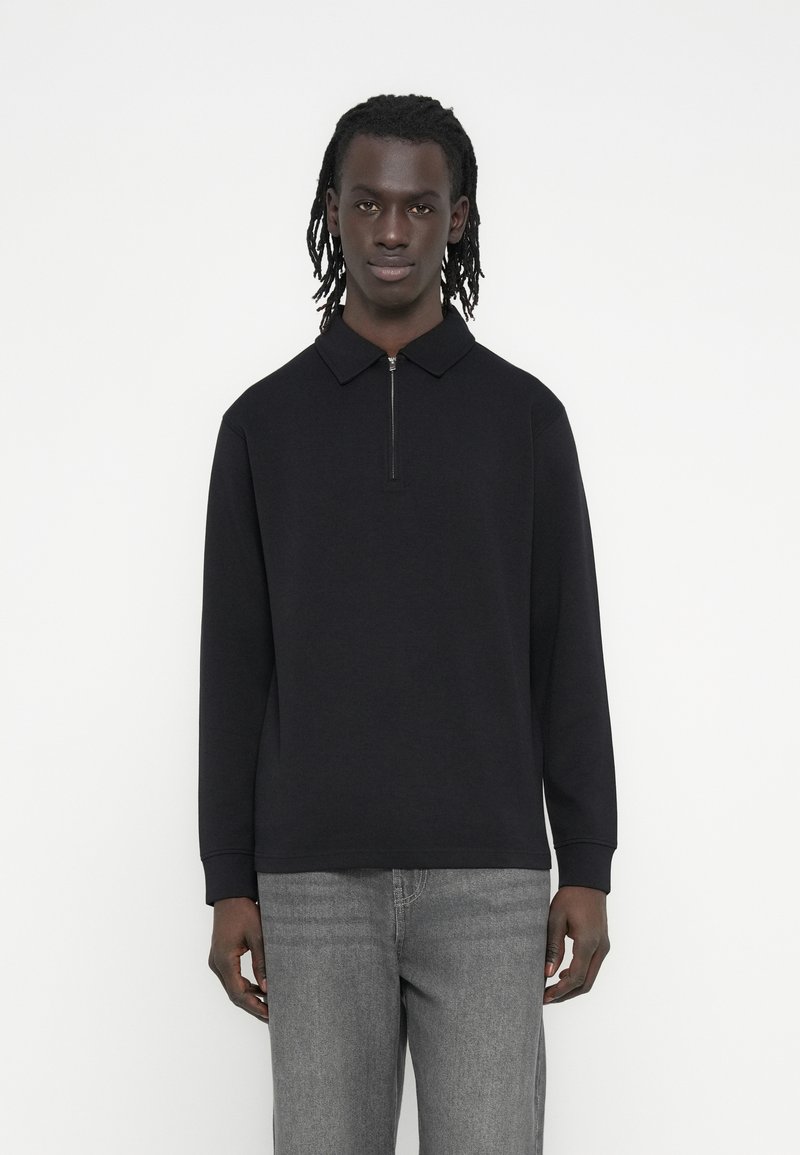 Μαύρο πουλόβερ με φερμουάρ στον γιακά και μακριά μανίκια, φτιαγμένο από λείο ύφασμα, συνδυασμένο με γκρι τζιν κανονικής γραμμής.