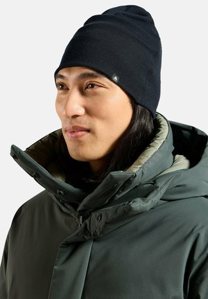 Man wearing black knit beanie and dark green winter jacket with high collar, looking to the right against a plain background.