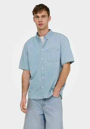 Junger Mann trägt hellblaues kurzärmeliges Jeanshemd und passende Jeans, steht mit einer Hand in der Tasche, neutraler Hintergrund.