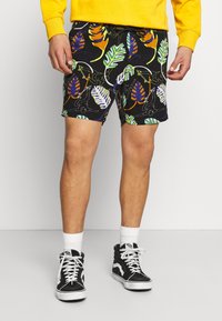 Shorts noirs avec des motifs de feuilles colorés en vert, orange et violet. Portés avec des baskets montantes noires et des chaussettes blanches.