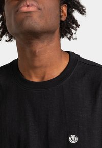 Czarna bawełniana koszulka z okrągłym dekoltem. Ma małe, białe logo haftowane na klatce piersiowej. Teksturowana tkanina z subtelnymi wariacjami.