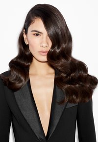 De longs cheveux noirs ondulés encadrent le visage. La silhouette porte un blazer noir ajusté avec un décolleté en V et un revers en satin.