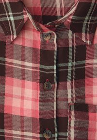 Gros plan sur une chemise à carreaux rouge et marron avec col, boutons et détail de poche cousue.