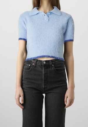 Personne portant un pull court duveteux bleu clair à manches courtes avec col et un jean taille haute foncé sur un fond uni.