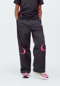 Osoba u crnim i ružičastim treniranim hlačama i usklađenoj crnoj majici s ružičastim detaljima stoji s rukama u džepovima.