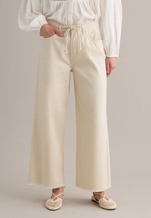 Vrouw draagt beige broek met hoge taille en wijde pijpen met trekkoorden in de taille, gecombineerd met een witte blouse met lange mouwen en beige geweven platte schoenen.