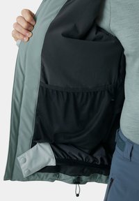 Het binnenwerk van de jas heeft een zwarte gaasvoering met een grijze stof aan de buitenkant. Inclusief drukknopsluiting en een verstelbare trekkoord aan de onderrand.