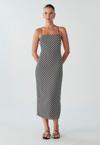 Midi šaty s čierno-bielym houndstooth vzorom, tenké ramienka, priliehavý strih a rovný silueta, v kombinácii s čiernymi sandálmi.