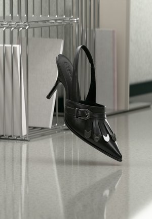 Pantof cu toc înalt, toc slingback, din piele neagră, cu vârf ascuțit, decorațiuni argintii în formă de pisică și curea cu cataramă, expus pe o suprafață reflectorizantă.
