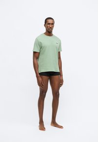 Πράσινο T-shirt με κοντά μανίκια φτιαγμένο από υφή υφάσματος, με στρογγυλή λαιμόκοψη και μικρή λεπτομέρεια λογότυπου στην αριστερή πλευρά, συνδυασμένο με μαύρο σορτς.