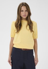 Gelbes, kurzärmeliges Strickoberteil mit geripptem Rundhalsausschnitt und weicher Textur. Getragen mit marineblauen Hosen und braunem Gürtel. Einfaches Design.