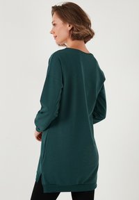 Törökzöld, hosszú ujjú pulóver, laza szabással, kerek nyakkal, oldalsó sliccekkel és bordázott mandzsetákkal. Puha tapintású, közepes hosszúságú forma.