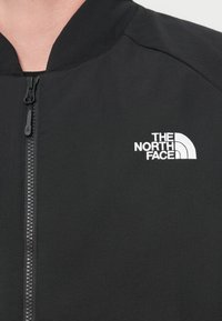 Μαύρο μπουφάν με ψηλό γιακά, με πλήρες φερμουάρ και λευκό κεντημένο λογότυπο στην αριστερή περιοχή του στήθους. Λείο υλικό, ελαφρύ υλικό.