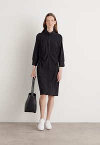 Zwarte jurk met een rits aan de voorkant, driekwart mouwen en ruches aan de zijkanten. Gepaard met een zwarte totebag en witte sneakers.