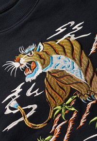 Sudadera negra con bordado detallado de tigre en dorado, naranja, azul y blanco. Incluye acentos verdes y motivos de relámpagos dinámicos.
