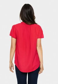 Blusa rossa a maniche corte con tessuto leggermente testurizzato, dettaglio arricciato sul retro e orlo arrotondato. Abbinata a jeans blu scuro.