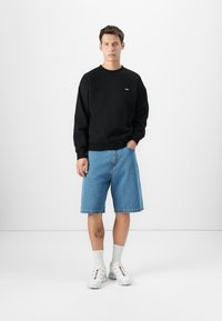 Črna športna majica, modri kavbojarski šort, in bele superge. Majica ima krožni ovratnik in logotip; šorti so do kolen s sproščenim fitom.