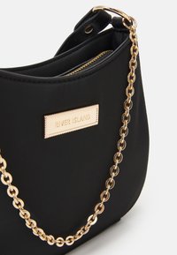 Czarna torebka o miękkiej fakturze, z złotym łańcuszkiem na ramię oraz metalową tabliczką z logo River Island. Zamek błyskawiczny na górze.