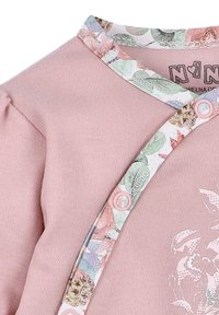 Lichtroze blouse met lange mouwen, voorzien van een kraagtrim met bloemenpatroon, roze drukknoopjes en een witte bloemenprint aan de voorkant. Zachte textuur.