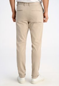 Man draagt beige slim-fit broek met achterzakken met rand en witte sneakers, staand tegen een witte achtergrond.