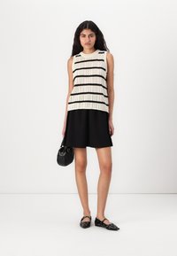Haut sans manches en tricot avec un motif rayé noir et crème, associé à une jupe noire et des ballerines noires, tenant un petit sac à main noir.