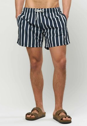 Mann trägt marineblaue und weiße vertikal gestreifte Badeshorts mit Kordelzugtaschen, kombiniert mit braunen Sandalen mit Schnalle, steht vor einem einfarbigen Hintergrund.