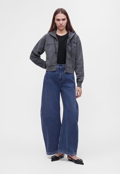 Femme portant un sweat à capuche court zippé gris foncé, un top noir, un jean large bleu et des ballerines pointues noires avec des détails de boucle argentée.