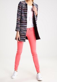 Abrigo largo texturizado multicolor con rayas, combinado con una camiseta gráfica blanca y jeans slim rosa brillante. Zapatillas blancas completan el atuendo.