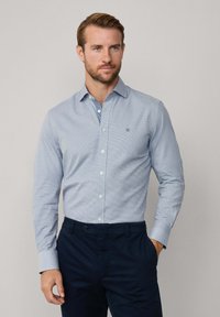 Muž so svetlým bradou, oblečený v svetlomodrej košeli s gombíkmi a tmavosivých nohaviciach, stojí na jednoliatom svetlom pozadí.