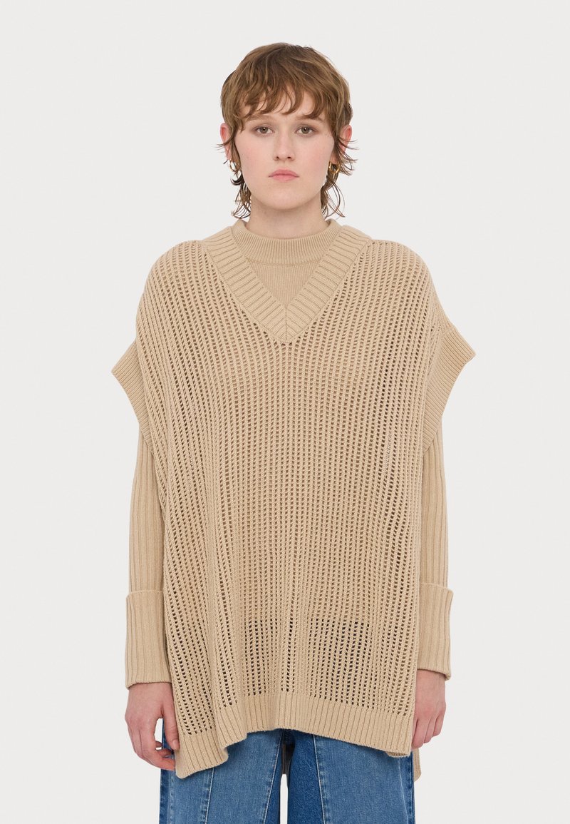 Beżowy, dziany oversize'owy poncho z dekoltem w serek, z otwartymi wzorami i ściągaczami na rękawach, w połączeniu z dopasowaną bluzką z długim rękawem.