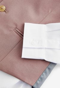 Tissu marron avec une texture lisse, présentant un détail de bouton. Une manche de chemise blanche affiche une finition texturée et une fermeture à bouton.