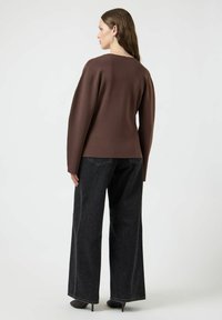 Brązowa bluzka z długim rękawem o luźnym kroju, zaprezentowana z czarnymi spodniami typu wide-leg i czarnymi szpilkami. Gładka faktura, minimalne szwy i prosty design.