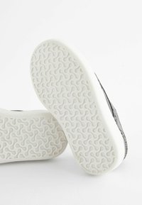 Zapatillas negras con textura, con una suela de goma blanca que presenta un diseño ondulado y ranuras para mayor tracción. El diseño de la suela enfatiza el agarre.