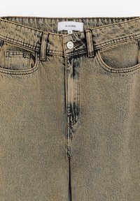 Jeans in denim beige con una trama sbiadita, dotati di una cintura, tasche frontali e chiusura con bottone argento. L'etichetta riporta "SCALPERS" e taglia 36.